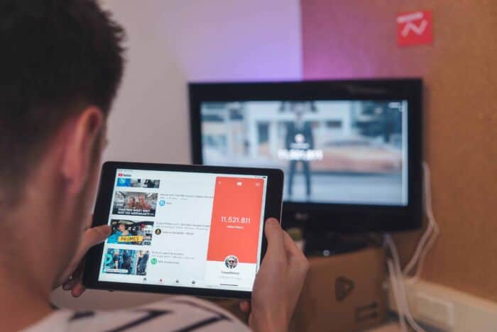 Back Of Person Looking At Youtube On An Ipad With Tv On Blurred In Background