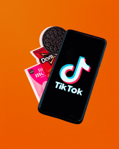 Phone With Tiktok Logo On The Screen And Different Food Brands Like Little Moons, Doritos And An Oreo Showing Out Of The Side