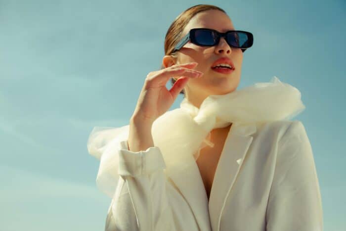 A Model Wearing A White Suit, Scarf, Black Sunglasses And A Slick Back Bun Stood Outside With Blue Sky Behind. Many Models Work With Brands On Fashion Influencer Marketing Campaigns.